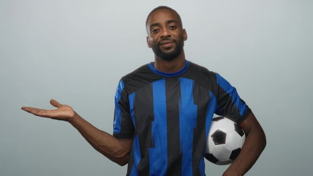 Man in blue and black soccer jersey holding a soccer ball under his arm with left palm up in studio against pale gray backdrop; confidence determination.