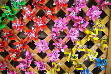 色鮮やかなビニール製の風車が無数に木製の格子状の壁に飾られている様子をクローズアップで捉えた写真です。赤ピンク黄色緑など様々な色が混ざり合っています。