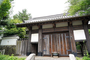 木々の緑に囲まれた重厚な木造の門をローアングルで捉えた写真です。瓦屋根と土壁そして石垣の組み合わせが日本の歴史的な寺院または城郭建築の風格を伝えています。