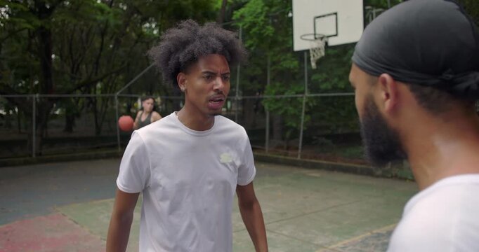 Two African American men arguing face to face on outdoor basketball court, confrontation escalating as one points angrily while the other listens tensely, emotional dispute