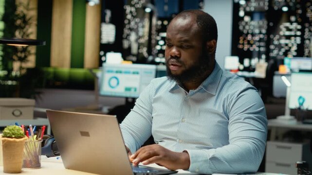 African american guy answering emails and documenting data from online insights for a multinational company, working after hours in a dark office. Enterprise objectives and development. Camera B.