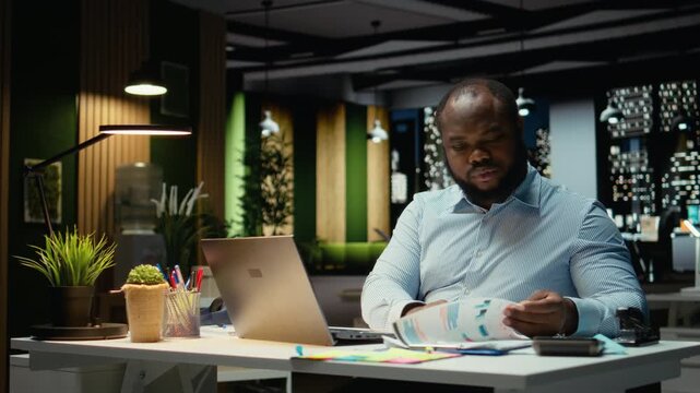 African american man does critical thinking and proofreading with papers before transcribing data on notebook, sitting in the office at night. Worker documenting important ideas. Camera A.