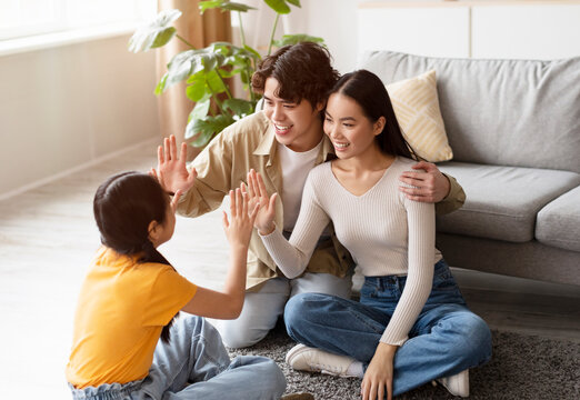 A young girl and two adults share a joyful moment in a comfortable living room. They engage in a hand game, smiling and encouraging each other, creating a warm atmosphere.