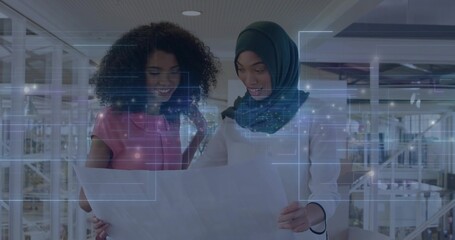 Two women wearing pink top and headscarf studying blueprint at office atrium with digital interface