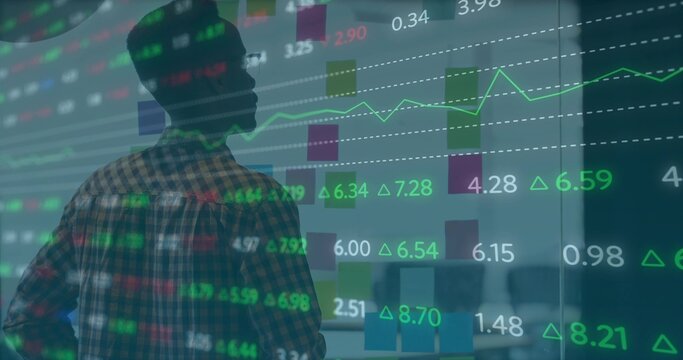 Studying analyst in plaid shirt reviewing stock charts on glass wall in office, with desk behind