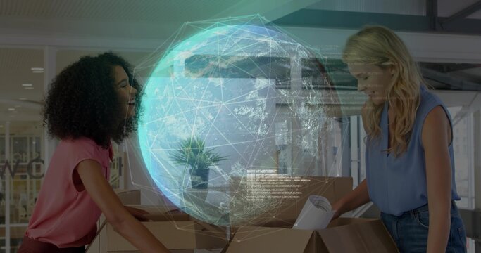 Unpacking two women in office wear reviewing holographic globe at office desk, with cardboard boxes