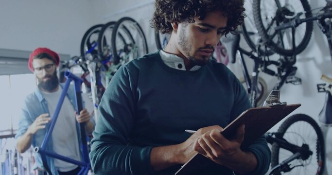 Curly-haired manager taking notes on clipboard inside bike workshop, with hanging frames and tools