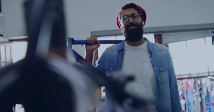 Bearded man with red beanie holding frame over shoulder inside bicycle workshop with repair stand