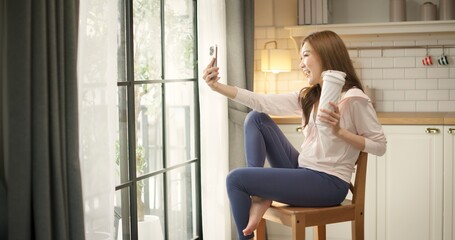 Obraz premium Young Asian woman in sportswear holds a protein shake bottle taking selfie with smartphone for social media after home workout. Healthy lifestyle, fitness motivation, self care, post-workout recovery.