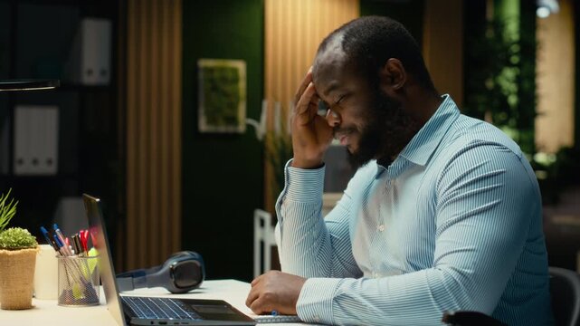 Black troubled worker rubbing his head in frustration and weariness, expressing fatigue and discomfort due to intense workload or failure. Angry frustrated man feels under pressure. Camera B.