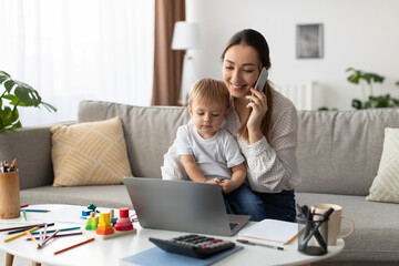 A woman is engaged in a phone call while seated on a couch. Her young child is sitting on her lap, playing with a laptop. Colorful toys and stationery are on the table nearby.