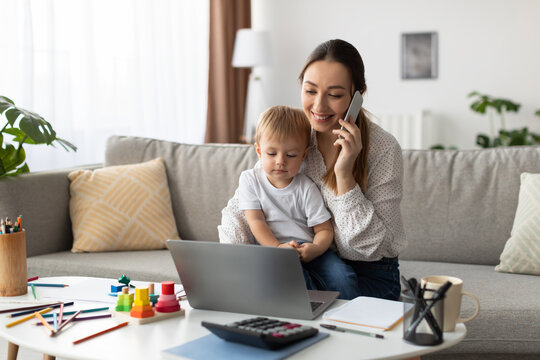 A woman is engaged in a phone call while seated on a couch. Her young child is sitting on her lap, playing with a laptop. Colorful toys and stationery are on the table nearby.