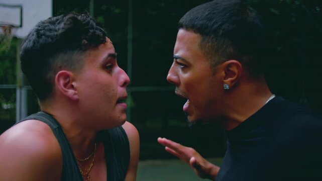 Two Hispanic men shouting face to face on basketball court, anger and frustration boiling over during argument, expressive gestures and emotional tension in daylight scene