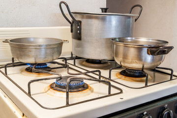 Three pots on active kitchen stove with visible blue flames, showcasing home cooking setup with aluminum and stainless steel cookware in rustic culinary environment