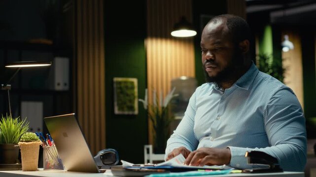 African american guy reviewing market data and building strategy for business goals, using spreadsheets on laptop for proofreading. Overtime after hours at multinational company. Camera B.