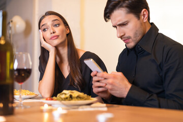 A couple sits at a table in a restaurant. The woman looks bored and disappointed while the man is focused on his phone. Their meals remain untouched, highlighting the disconnection between them.