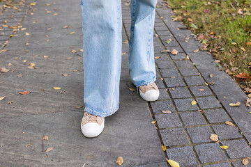 Person wearing light blue jeans and beige sneakers standing on a leaf-covered pathway in a park