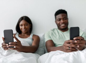 A man and woman sit together in bed, both focused on their smartphones. They appear relaxed and engaged, enjoying their time together in a calm atmosphere.
