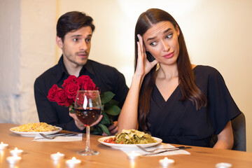 A man offers a bouquet of red roses to a woman during a candlelit dinner. She looks skeptical or unhappy, while plates of food and wine glasses sit on the table.
