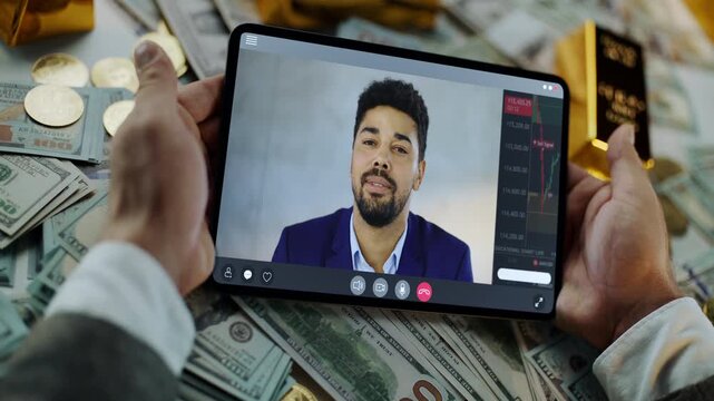 Businessman hands holding a tablet during a video conference with a financial advisor, analyzing the stock market trends over a desk covered with money, gold bars, and cryptocurrency coins