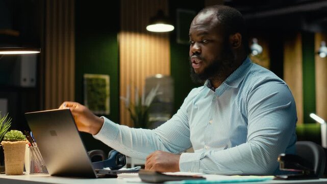 Black male associate answering online video call in office late at night, reports data for new project and engaging in transparent discussion. Exchanges ideas to strengthen relations. Camera B.