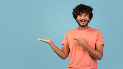 A smiling young man with curly hair and a beard is casually pointing at something on his palm. He is in a blue studio, creating a friendly and inviting atmosphere for advertising a product.