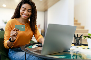 A woman sits at her desk in a bright workspace, happily making a purchase online. She holds a...