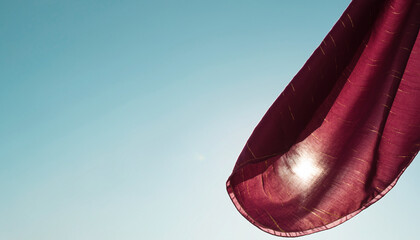 A silk scarf corner fluttering mid-air, rich burgundy with subtle gold stitching, suspended against a pale blue sky gradient; scarf anchored upper-right, 70% open sky to the left for text overlays. Na