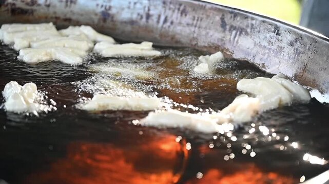 street food deep fried dough vendor hot oil outdoor market ladle golden crispy snack breakfast slow motion video capture shows fresh dough lifted from bubbling oil with steam and slight drizzle evokin