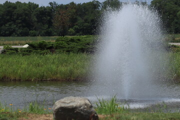 Small Fountain with Water Splashes and Droplets against Green Grass and Trees