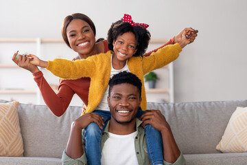 A joyful family shares a moment in their living room. The young girl sits on her father's shoulders...