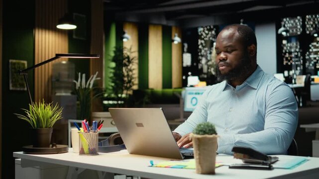 Black employee reading emails and documenting data from web insights for a multinational company, working after hours in a dark office. Enterprise objectives and development. Camera A.