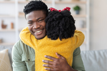A father and daughter enjoy a heartfelt hug in their living room. The girl is wearing a red bow,...