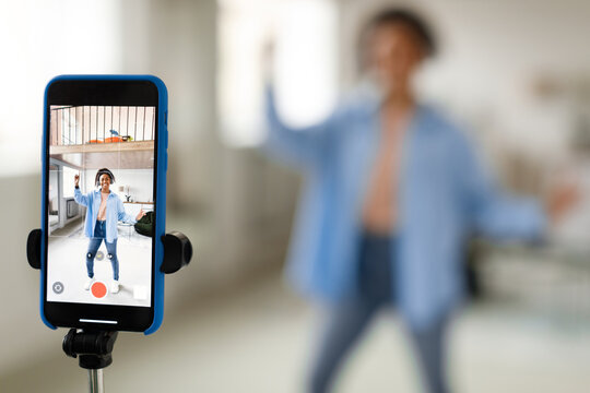 A young individual is energetically performing a dance routine in a well-lit room, with a smartphone set on a tripod capturing the moves. The person appears focused and lively.