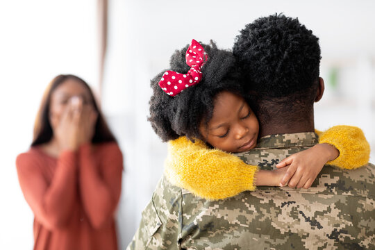 A soldier holds his young daughter close as she rests her head on his shoulder. A woman in the background wipes tears of joy, celebrating their heartfelt reunion at home.