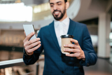 A man in a blue suit stands in a contemporary office space. He holds a coffee cup in one hand and a smartphone in the other, looking at the screen with a smile.