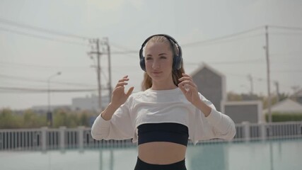 Confident young female athlete wearing headphone before warm up exercise near swimming pool in the morning. Fitness, focus, and the use of music for motivation during an active lifestyle routine.