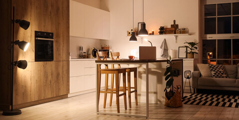 Interior of modern kitchen with counters, table and glowing lamps at night