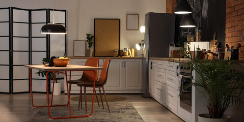 Interior of stylish kitchen with counters, dining table and glowing lamps at night