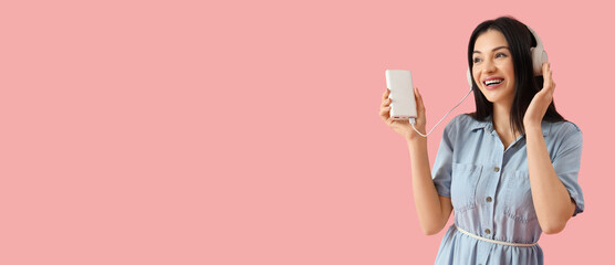 Beautiful young woman with modern power bank and headphones on pink background