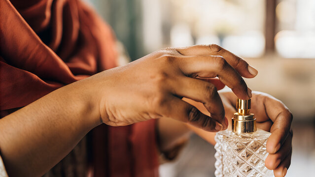 Indian Muslim woman&rsquo;s hand (warm brown skin) applying attar from a crystal bottle with gold cap onto inner wrist. Blurred deep red dupatta in background, soft natural light.