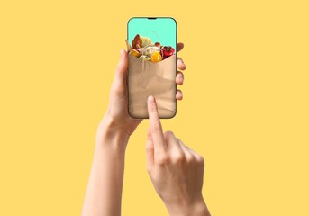 Hands holding mobile phone with grocery bag popping out of screen on yellow background
