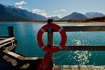 A bright orange life preserver hangs on a wooden jetty railing in Glenorchy, New Zealand on May 18, 2025. The turquoise lake and majestic, snow-capped mountains form a stunning, sunlit backdrop.