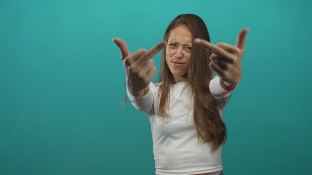 Woman showing middle fingers to camera in teal studio with extended fingers and scrunched face; bold defiance.