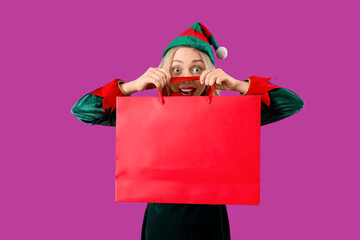 Happy young woman dressed as elf with shopping bag on purple background