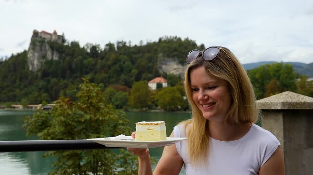 Smiling woman shaking famous Bled cream cake dessert outdoors at Kavarna Park Cafe, medium static