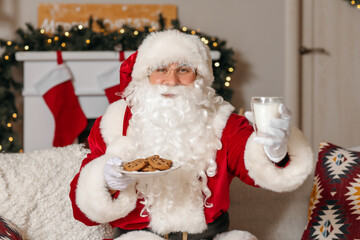 Santa Claus with cookies and glass of milk sitting on sofa at home