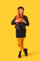 Beautiful young happy woman in autumn outfit with pumpkin on yellow background