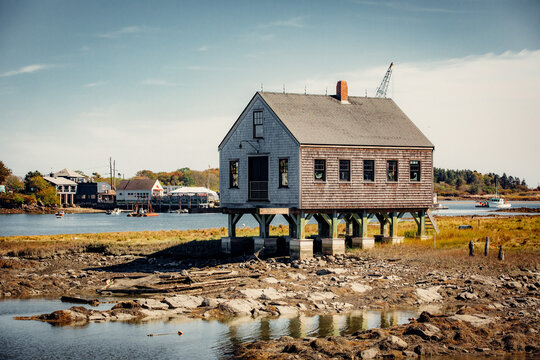 Historic wooden boathouse standing on stilts along the rocky coast during low tide. Scenic view of a coastal village with fishing boats and calm water under a clear blue sky.  - Powered by Adobe