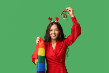 Young Asian woman in Christmas reindeer horns with LGBT flag and mistletoe on green background